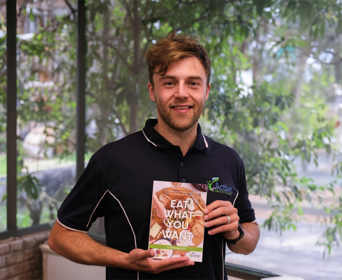 Luke Hassan from Active Health Clinic holding his book titled 'Eat What You Want' and smiling.