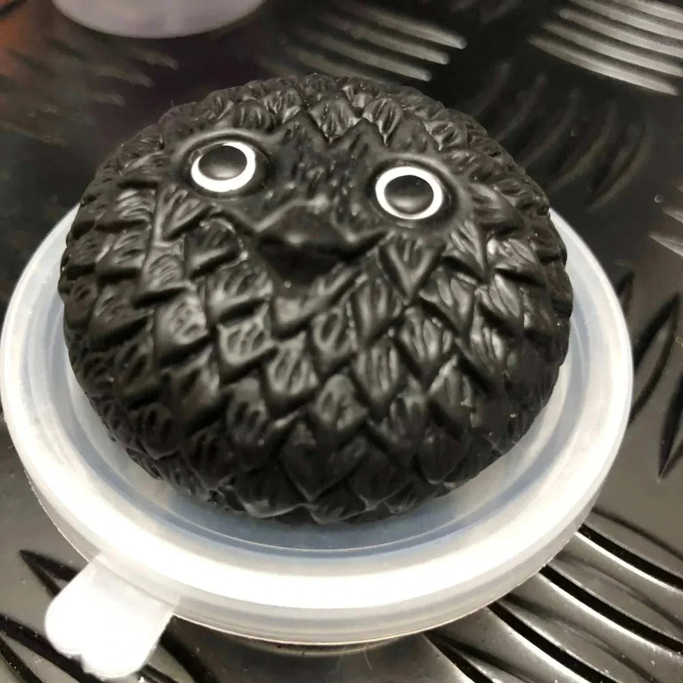Black Jelly Mochi Squishy Animal toy with textured surface and large eyes on a white container lid