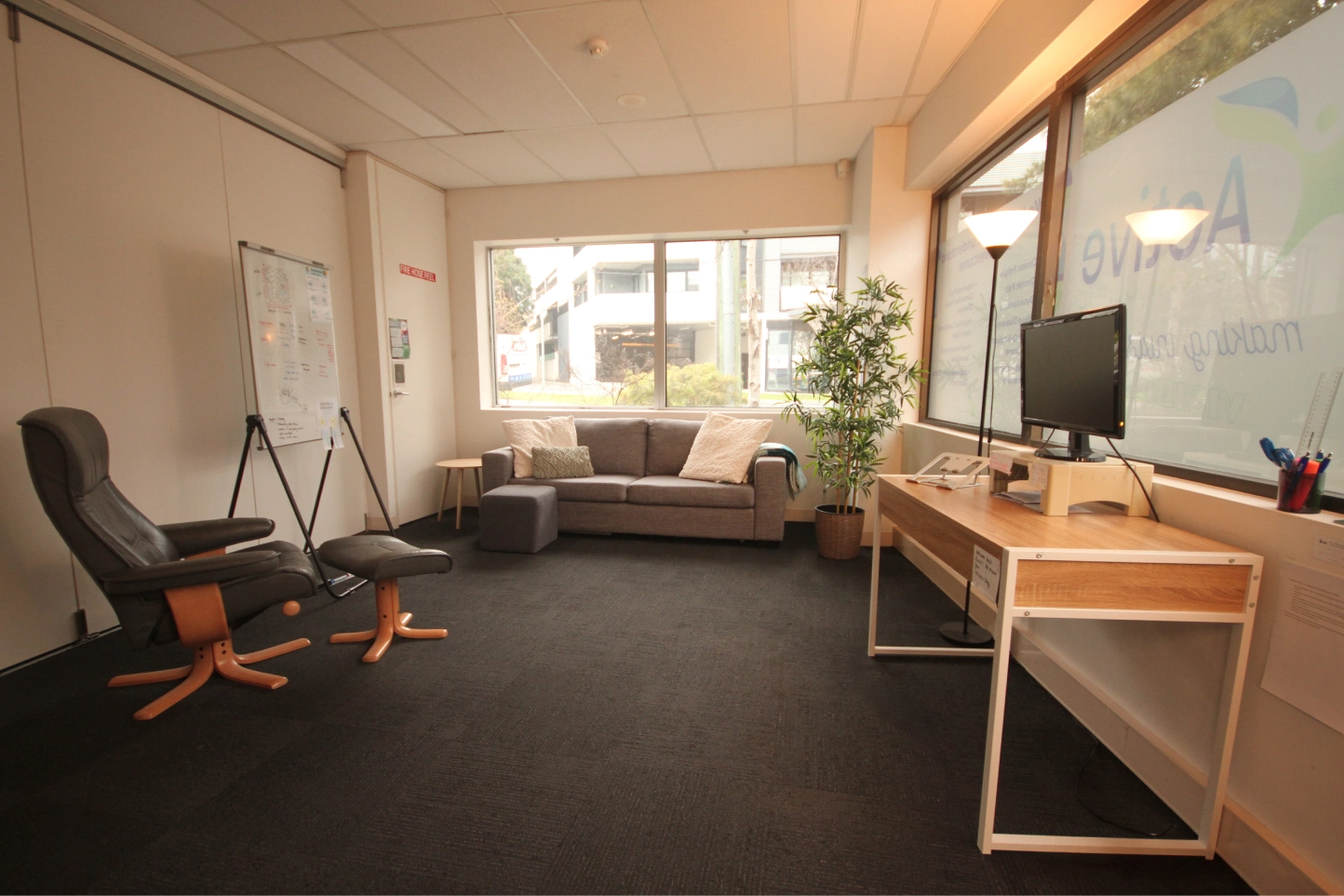 Comfortable consultation room at an allied health clinic with desk, computer, sofa, whiteboard, and natural daylight.